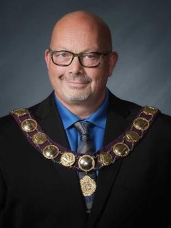 Mayor Patrick Van Minsel headshot. There is a man wearing a suit, glasses and a ceremonial necklace.